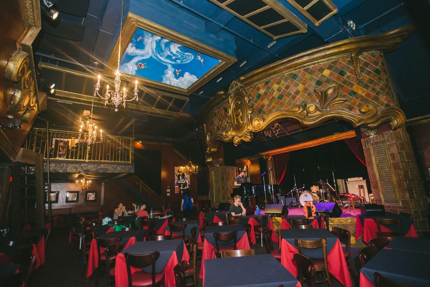 A dimly lit bar with a stage, tables, and red tablecloths.